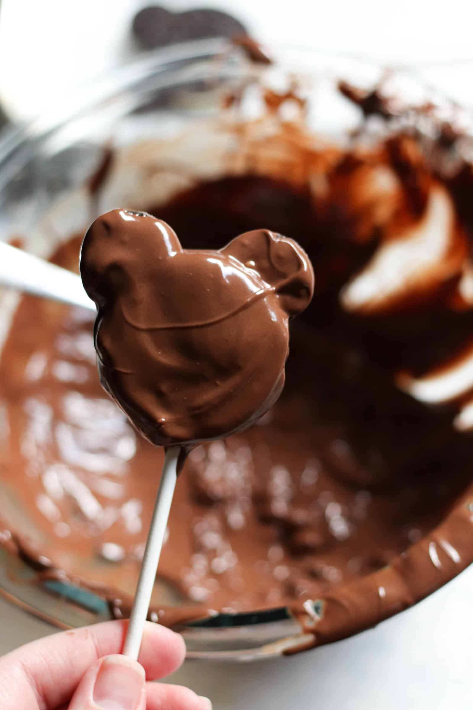 Spooning melted chocolate on top of Oreos on a stick shaped like Mickey Mouse. A bowl of melted chocolate in the background.