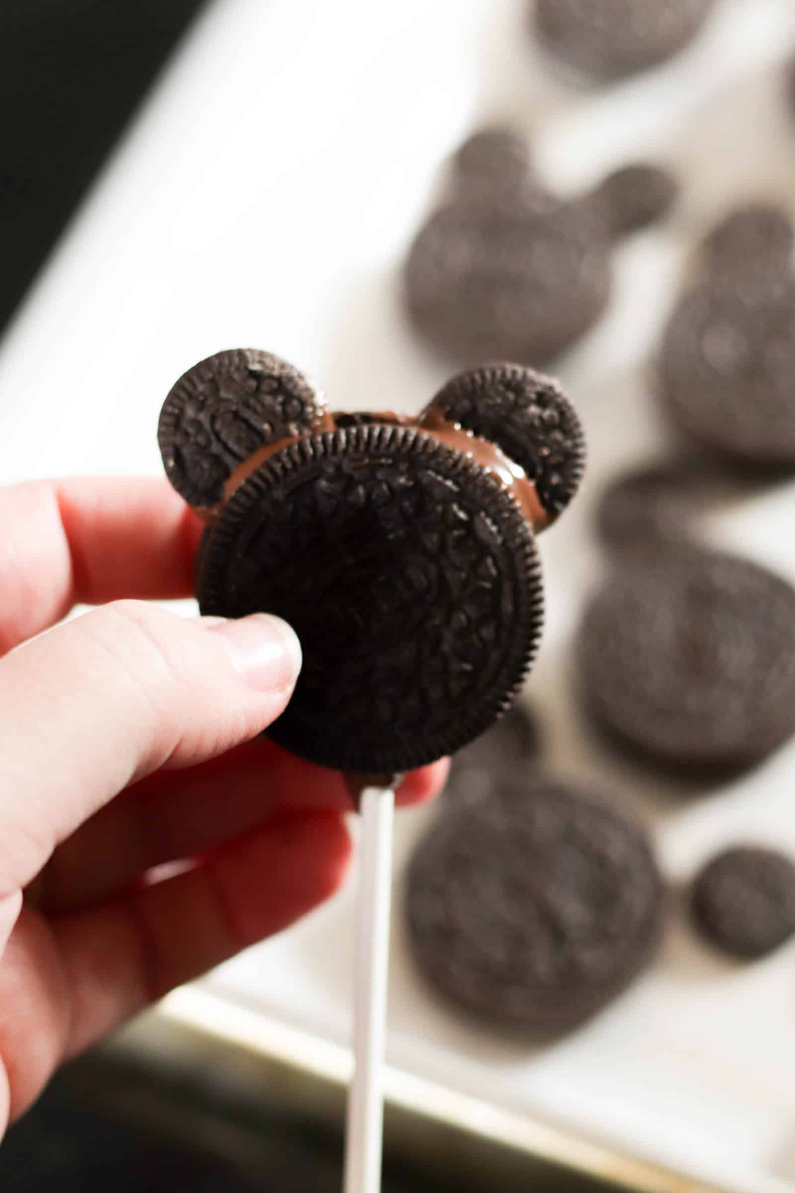 A hand holding an oreo on a stick with two mini oreos added on top to create ears. Other Oreos on a sheet pan in the background.