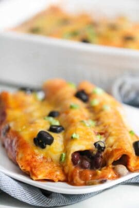 Close-up of cheesy enchiladas topped with black olives and green onions, served on a white plate with a dish in the background.