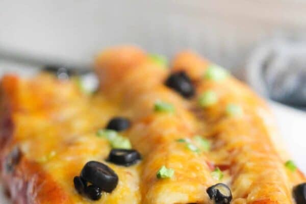 Close-up of cheesy enchiladas topped with black olives and green onions, served on a white plate with a dish in the background.