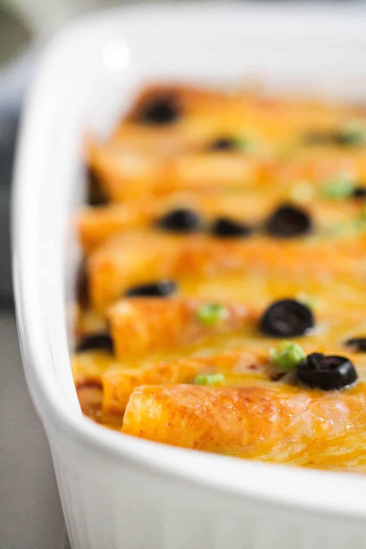 Close-up of a casserole dish filled with baked enchiladas topped with melted cheese, black olives, and green onions.