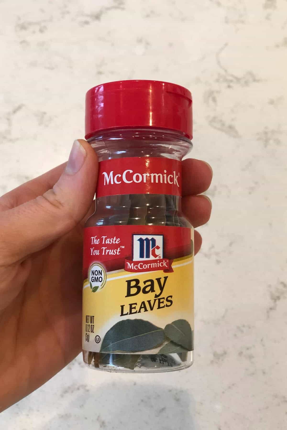 Hand holding a small jar of McCormick Bay Leaves, labeled "Non-GMO" and "The Taste You Trust," with a red cap and yellow label.