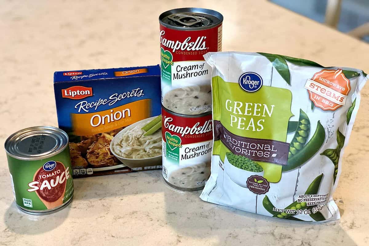 A box of Lipton onion soup mix, two cans of Campbell's cream of mushroom soup, a small can of Kroger tomato sauce, and a bag of Kroger frozen green peas on a kitchen counter.