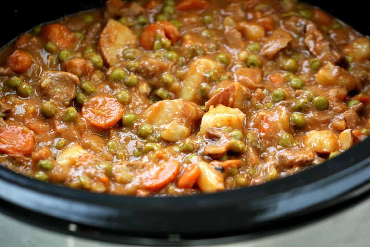 A stew with chunks of meat, carrots, potatoes, and peas in a thick sauce cooked in a slow cooker.