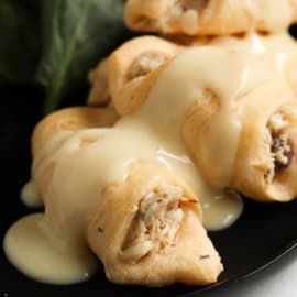 Close-up of stuffed pastry rolls drizzled with a creamy white sauce on a black plate, accompanied by fresh spinach leaves.