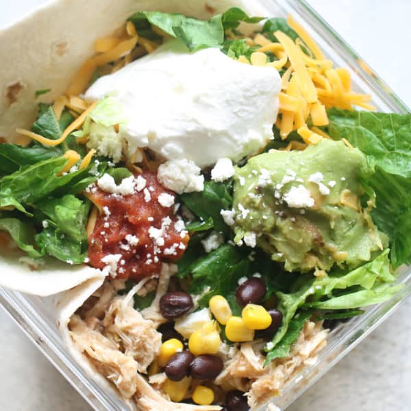 A glass container holds a tortilla, shredded cheese, sour cream, guacamole, salsa, lettuce, shredded chicken, black beans, and corn, arranged as a deconstructed taco salad.