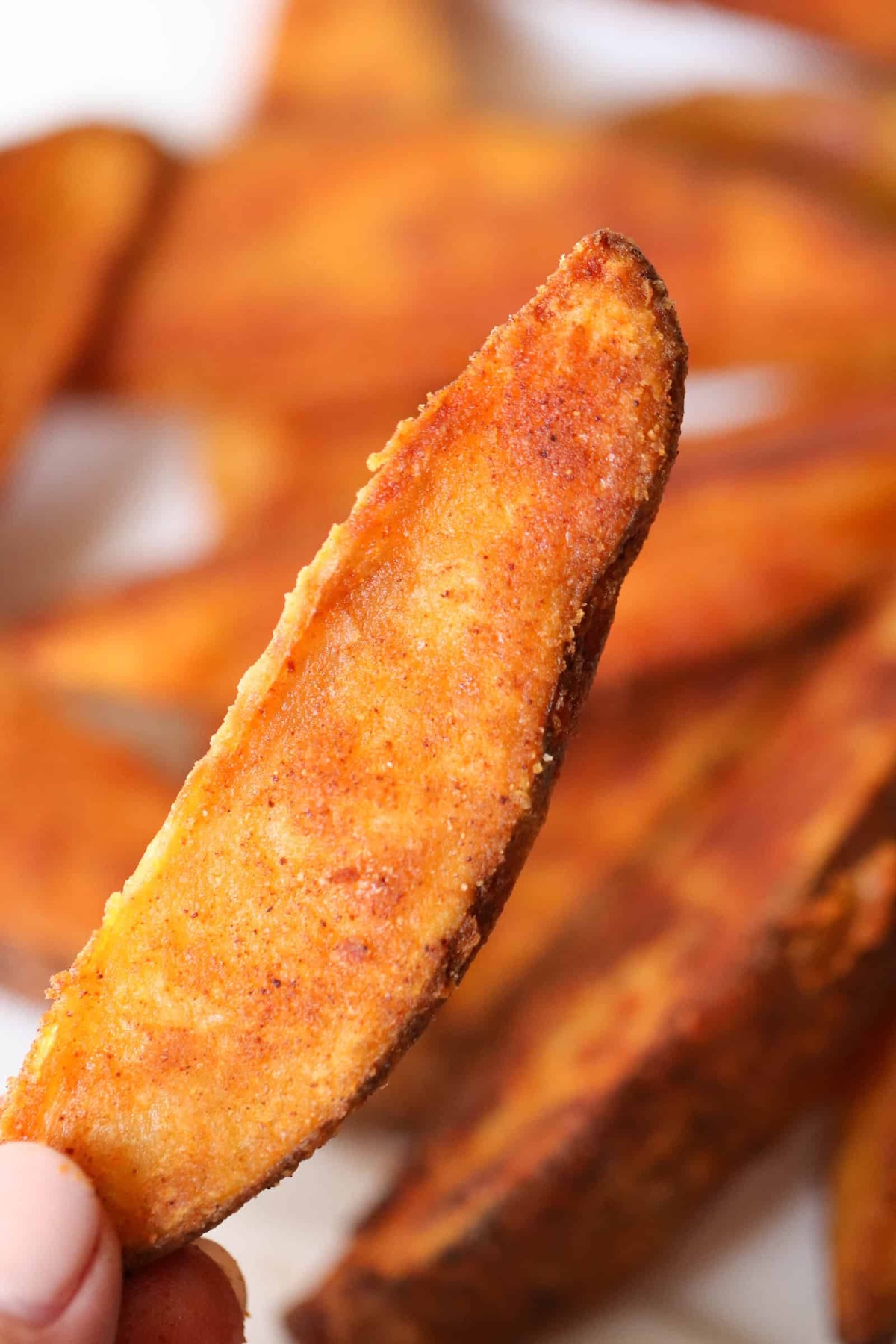 Close-up of a hand holding a seasoned potato wedge with a crispy golden-brown crust. More potato wedges are blurred in the background.