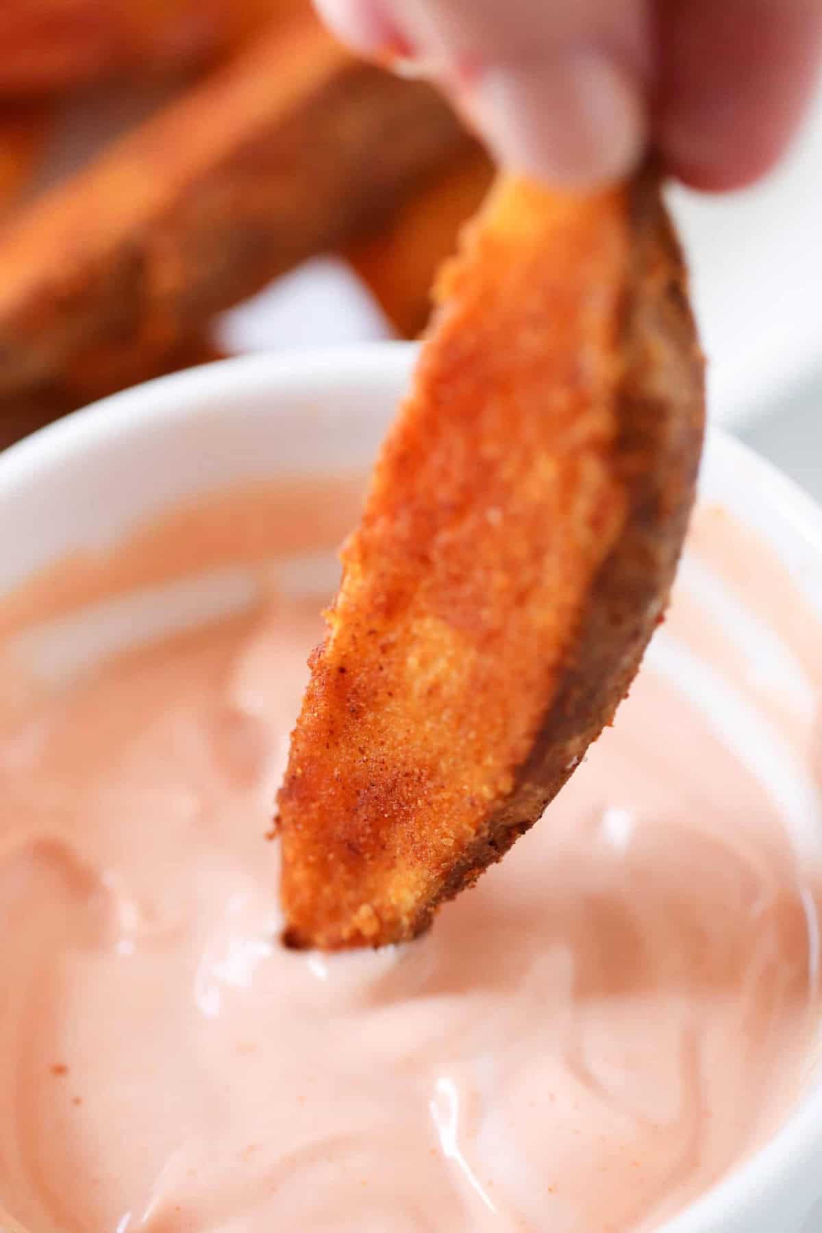 A hand dips a seasoned potato wedge into a creamy dipping sauce.