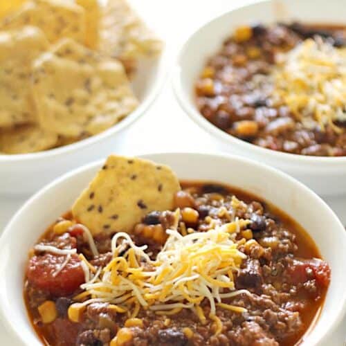 Three bowls are arranged with two containing chili topped with shredded cheese and one with tortilla chips.