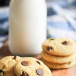 A bottle of milk stands next to a stack of three chocolate chip cookies with additional cookies scattered nearby on a wooden surface.