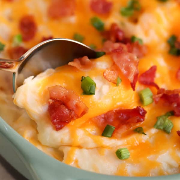A close-up of mashed potatoes topped with melted cheese, bacon pieces, and chopped green onions, with a spoon scooping a portion.