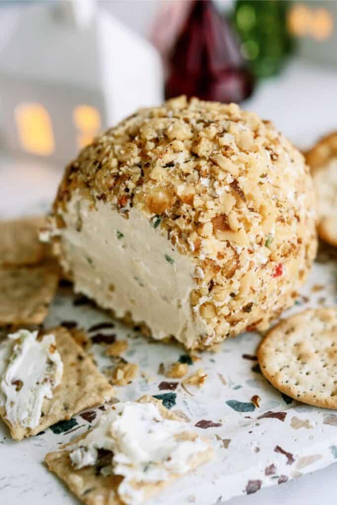 A cheese ball coated with crushed nuts, partially sliced and surrounded by assorted crackers, is displayed on a white surface.