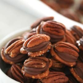 A white bowl is filled with pecan halves sandwiching chocolate, with more of the same in the background.