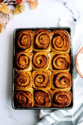 A tray of twelve evenly spaced, baked pumpkin cinnamon rolls placed on a white surface with scattered leaves and a striped cloth nearby.