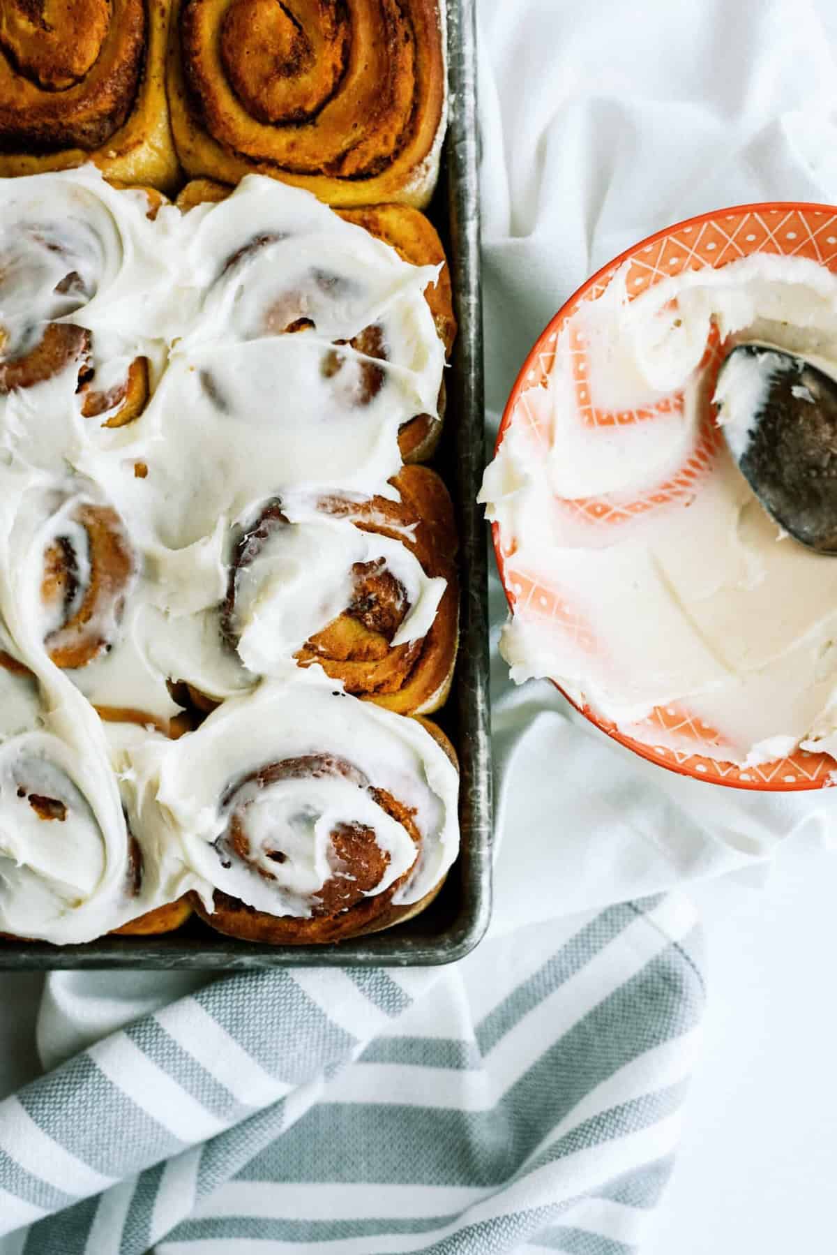 A tray of pumpkin cinnamon rolls partly covered with white frosting is next to an orange bowl containing more frosting and a spoon. A white cloth with gray stripes lies beneath.