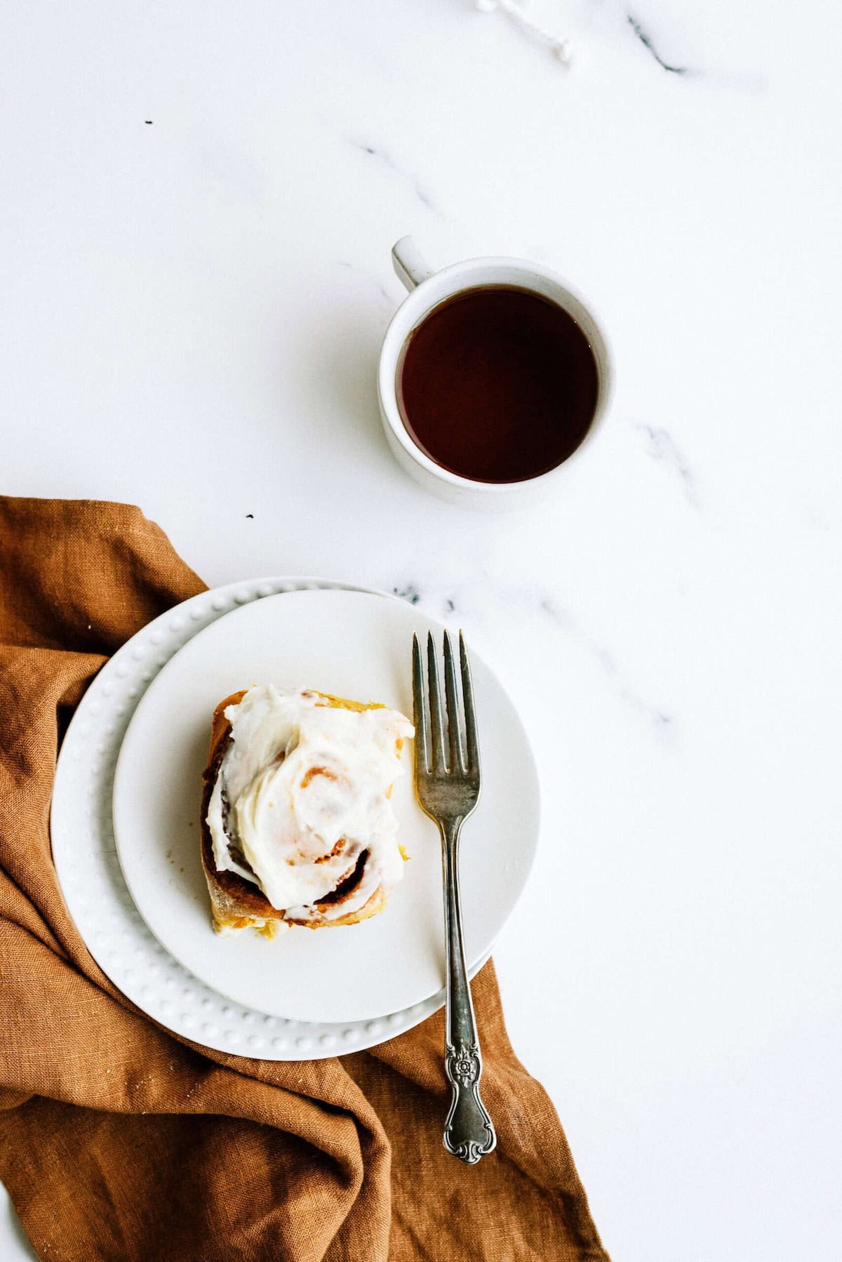 A cinnamon roll with white frosting on a white plate, accompanied by a fork, a brown napkin, and a cup of black coffee on a white marble surface.