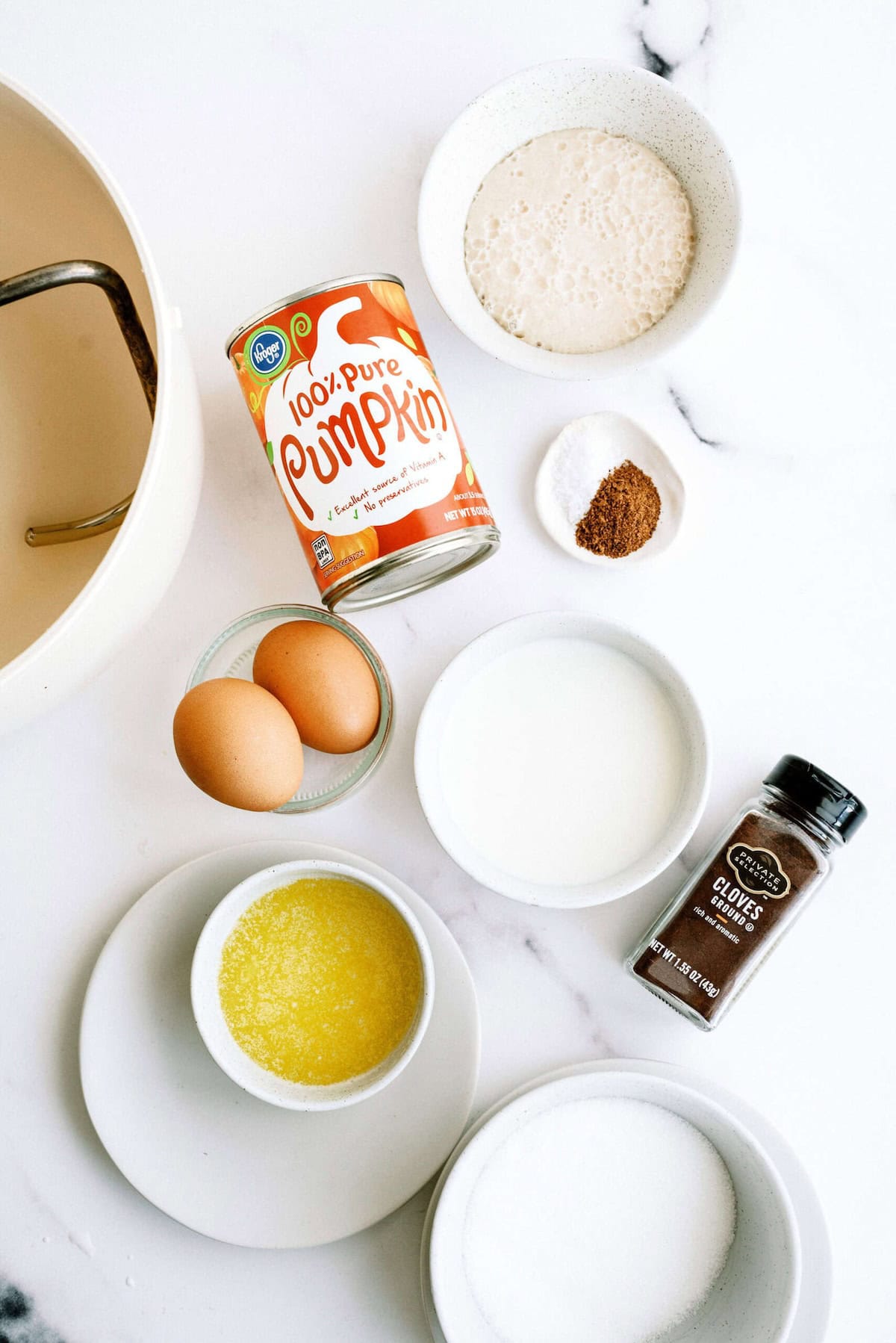 Ingredients for a recipe are laid out, including a can of pumpkin puree, two eggs, melted butter, spices, sugar, milk, and a small bowl of flour.