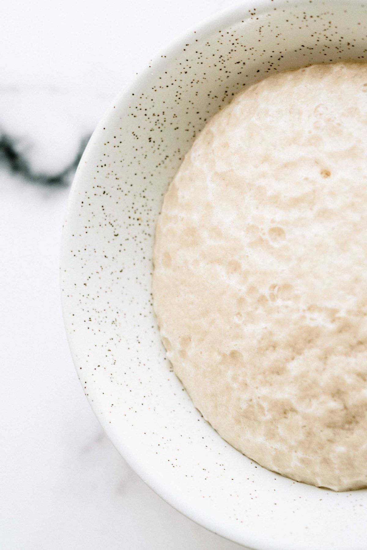 Overhead view of bread dough being mixed in a white stand mixer bowl. The dough is partially covering the metal mixing attachment.