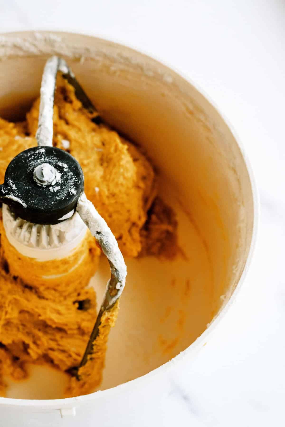 Overhead view of bread dough being mixed in a white stand mixer bowl. The dough is partially covering the metal mixing attachment.