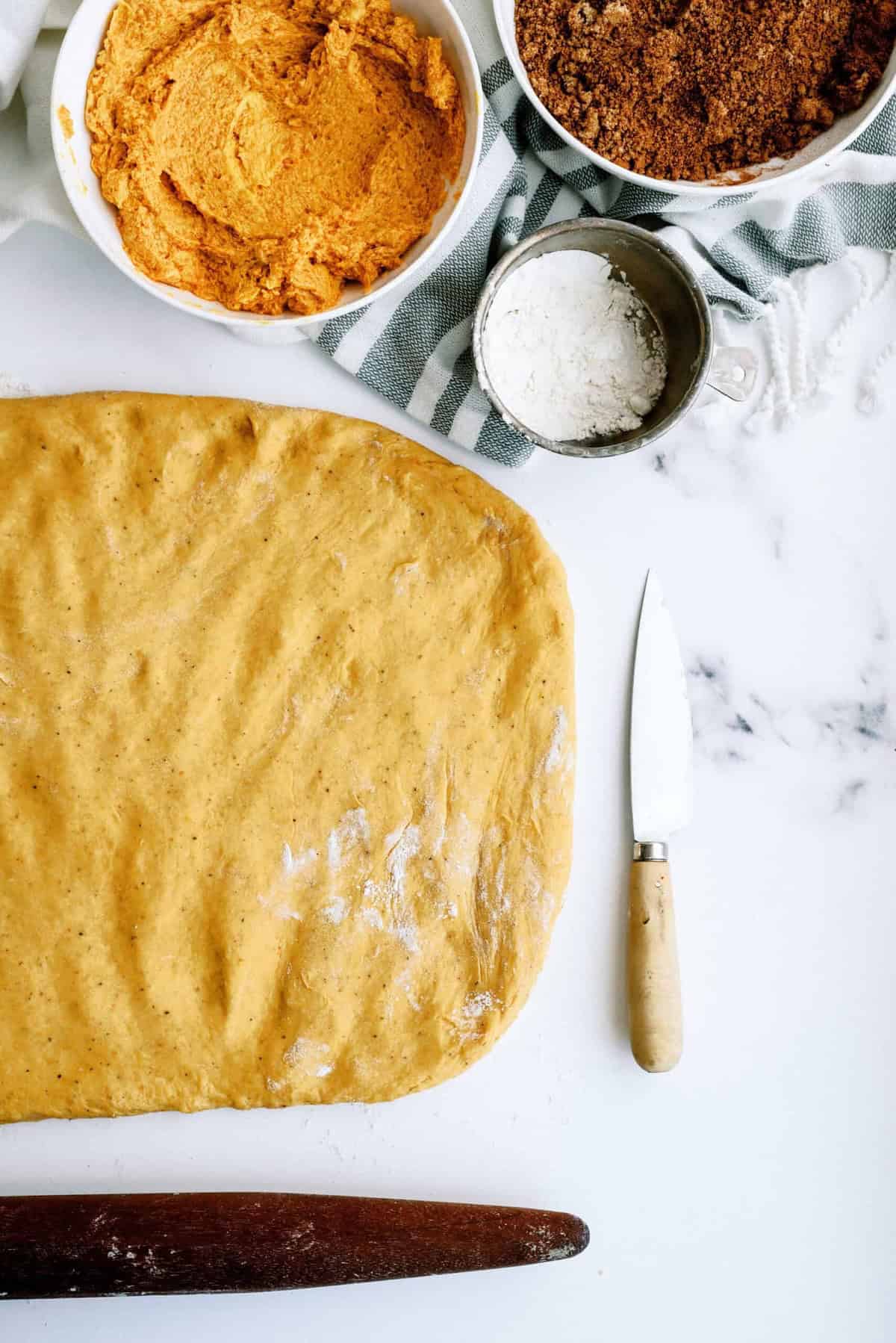 A flat rectangle of yellow dough on a white surface beside a rolling pin, knife, bowls of flour, pumpkin mix, and brown sugar with a green-striped cloth in the background.