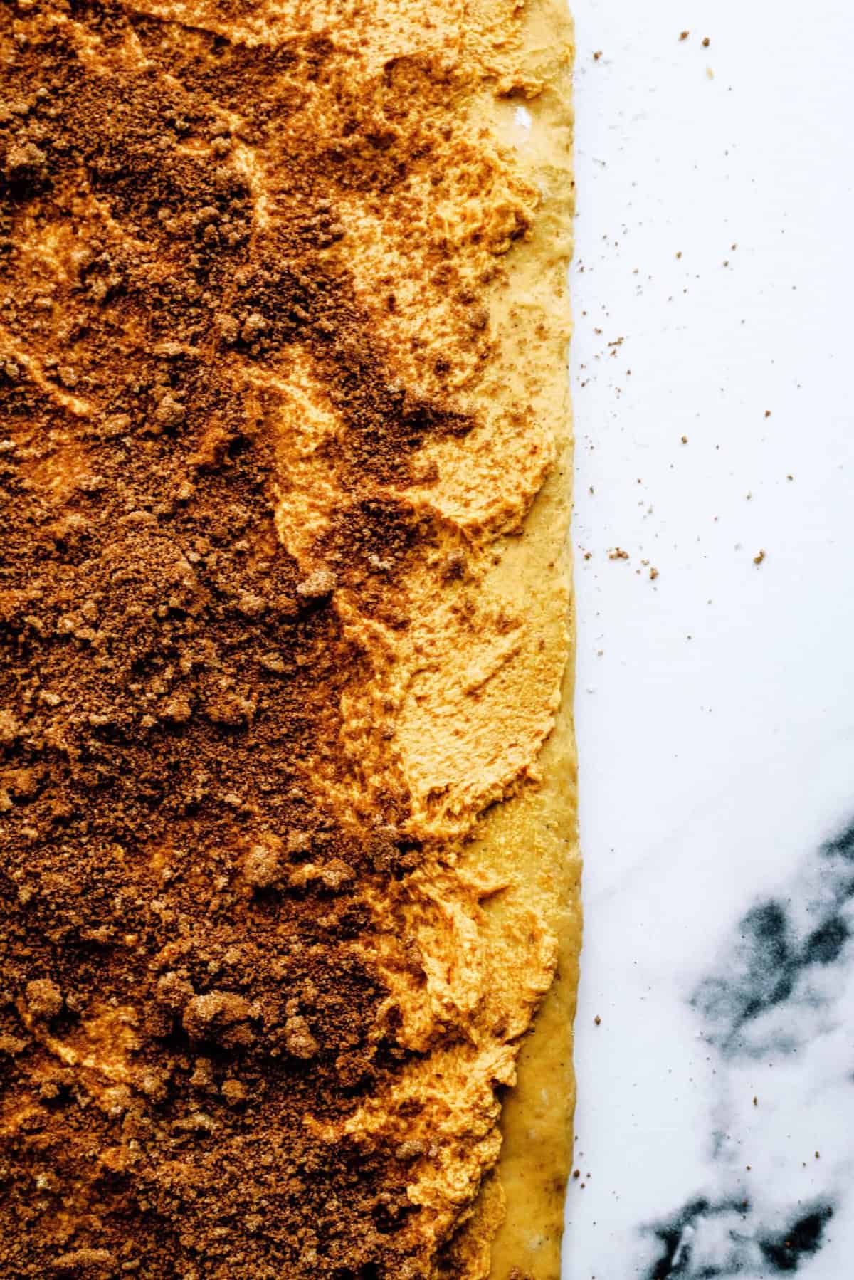 Close-up of rolled-out dough covered in a layer of brown sugar and cinnamon mixture on a white and marble surface.