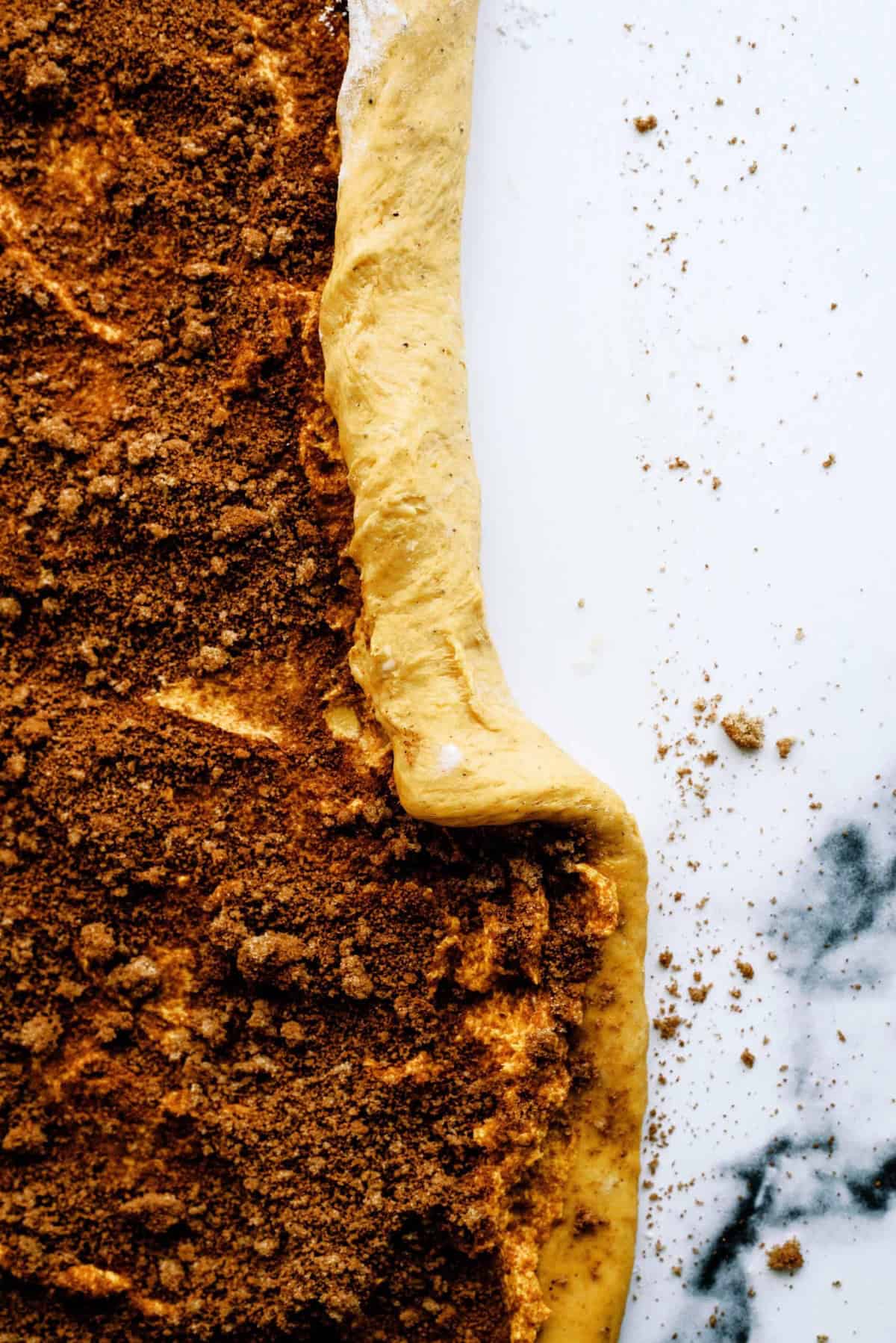 Close-up of partially rolled dough covered with a cinnamon and sugar mixture on a white, marbled surface.