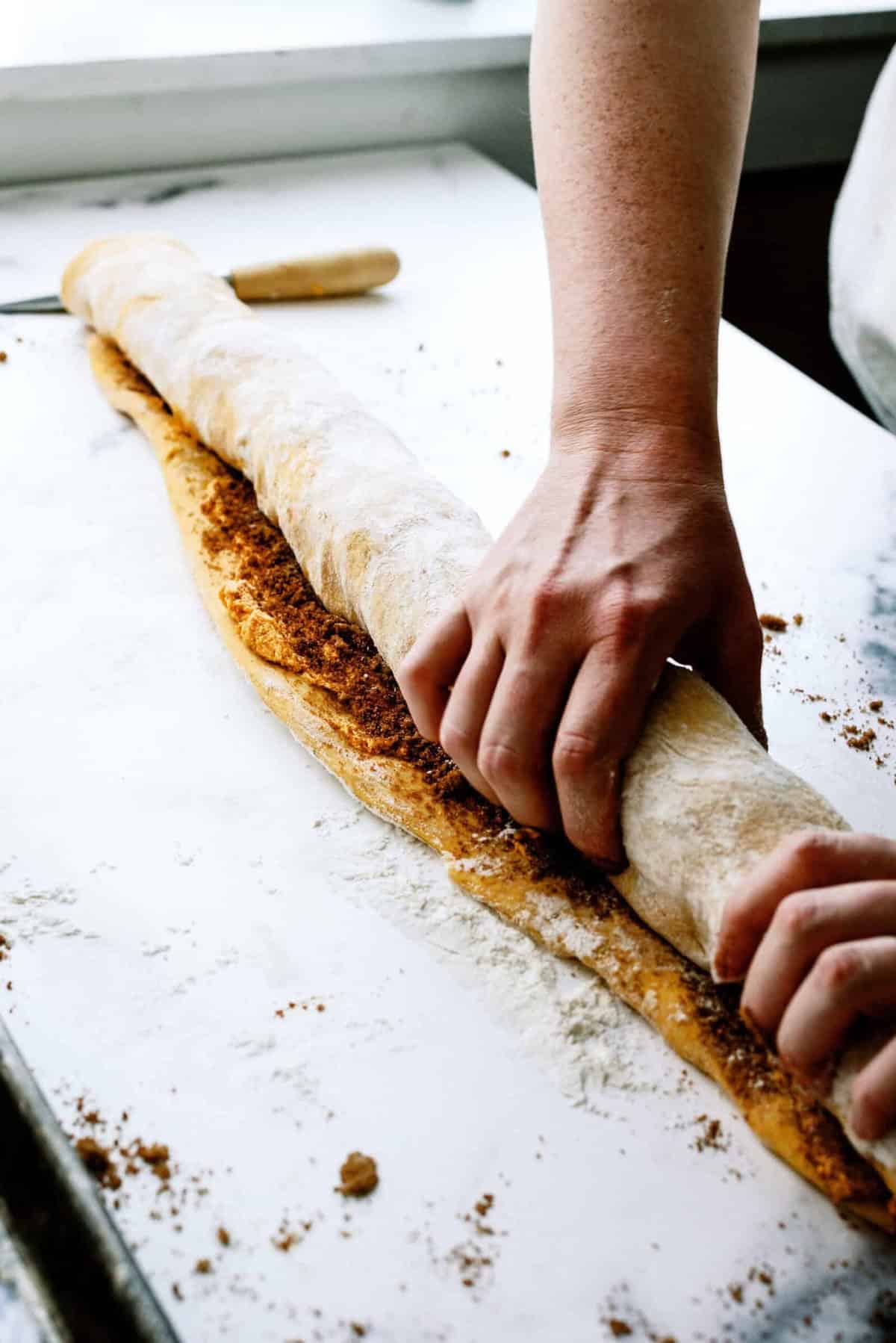 A person rolls dough filled with a brown sugar and cinnamon mixture on a floured surface. A rolling pin and baking tray are visible in the background.