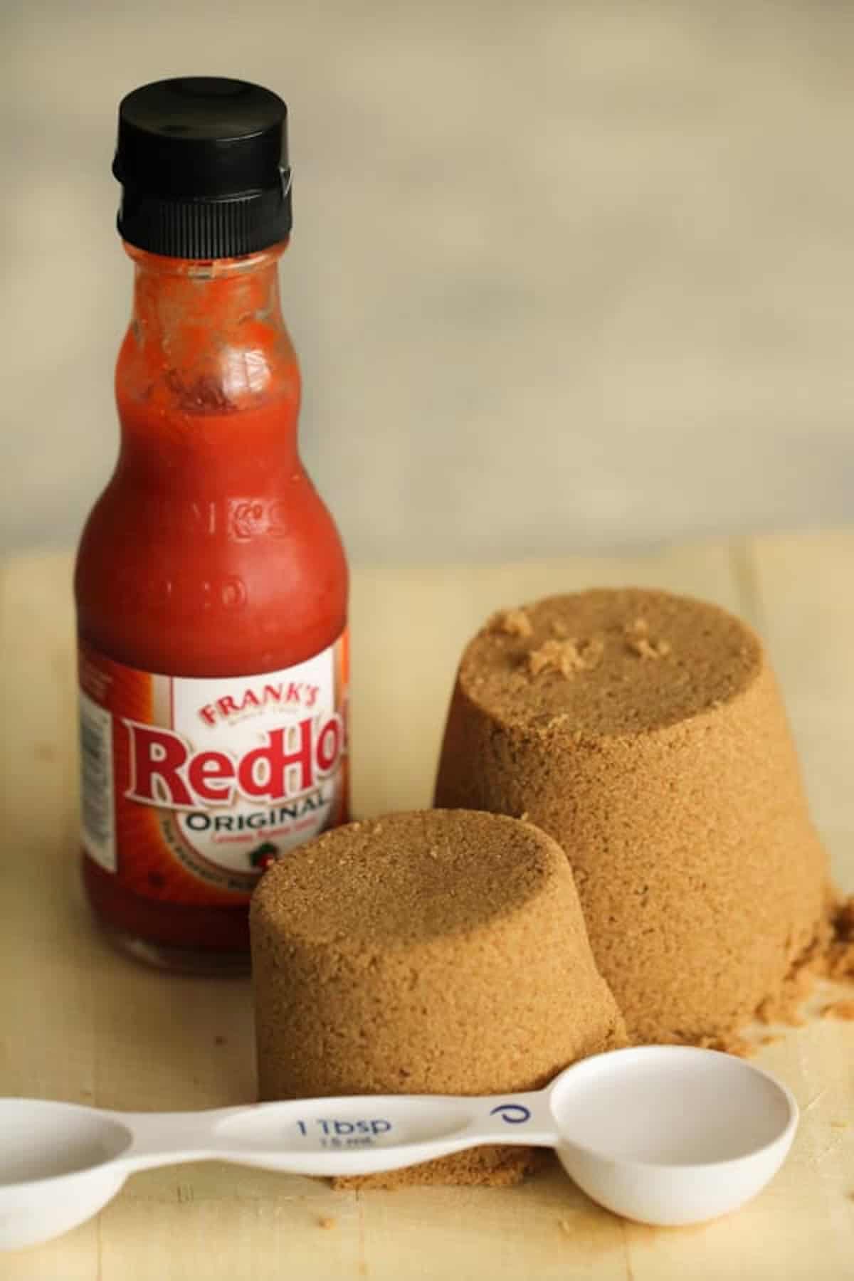 A bottle of Frank's RedHot Original sauce stands next to two packed mounds of brown sugar and two white measuring spoons.