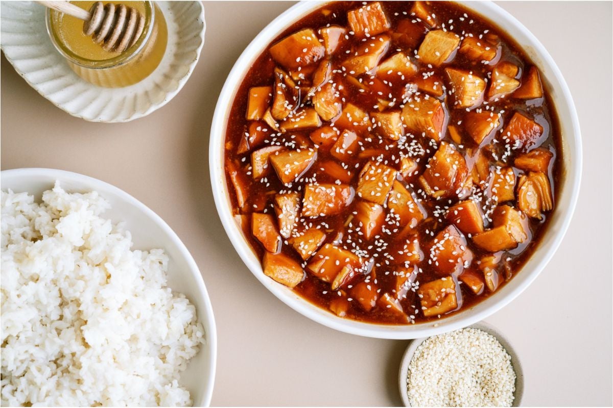 A serving bowl of Slow Cooker Honey Sesame Chicken topped with sesame seeds, a bowl of rice  and two small bowls of honey and sesame seeds.