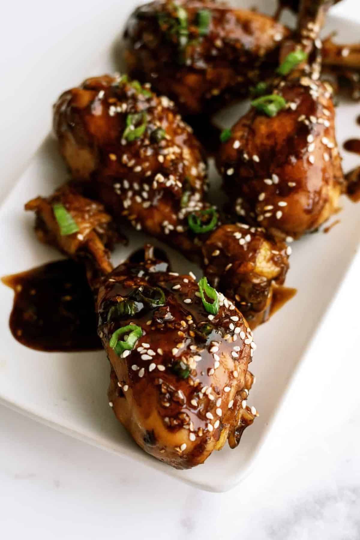 A plate of glazed chicken drumsticks garnished with sesame seeds and chopped green onions.