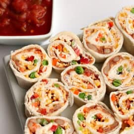 A plate of neatly arranged, colorful pinwheel wraps filled with various vegetables and a side of tomato-based dip in a square white dish.