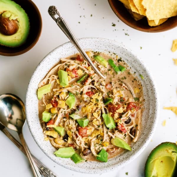 A bowl of chicken tortilla soup topped with diced avocado and surrounded by two spoons and a bowl of tortilla chips. An avocado half and scattered herbs are visible in the background.