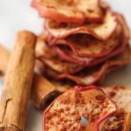 A stack of dried apple slices with cinnamon, accompanied by a cinnamon stick, placed on a white surface.