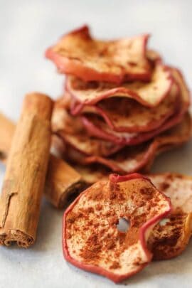 A stack of dried apple slices with cinnamon, accompanied by a cinnamon stick, placed on a white surface.