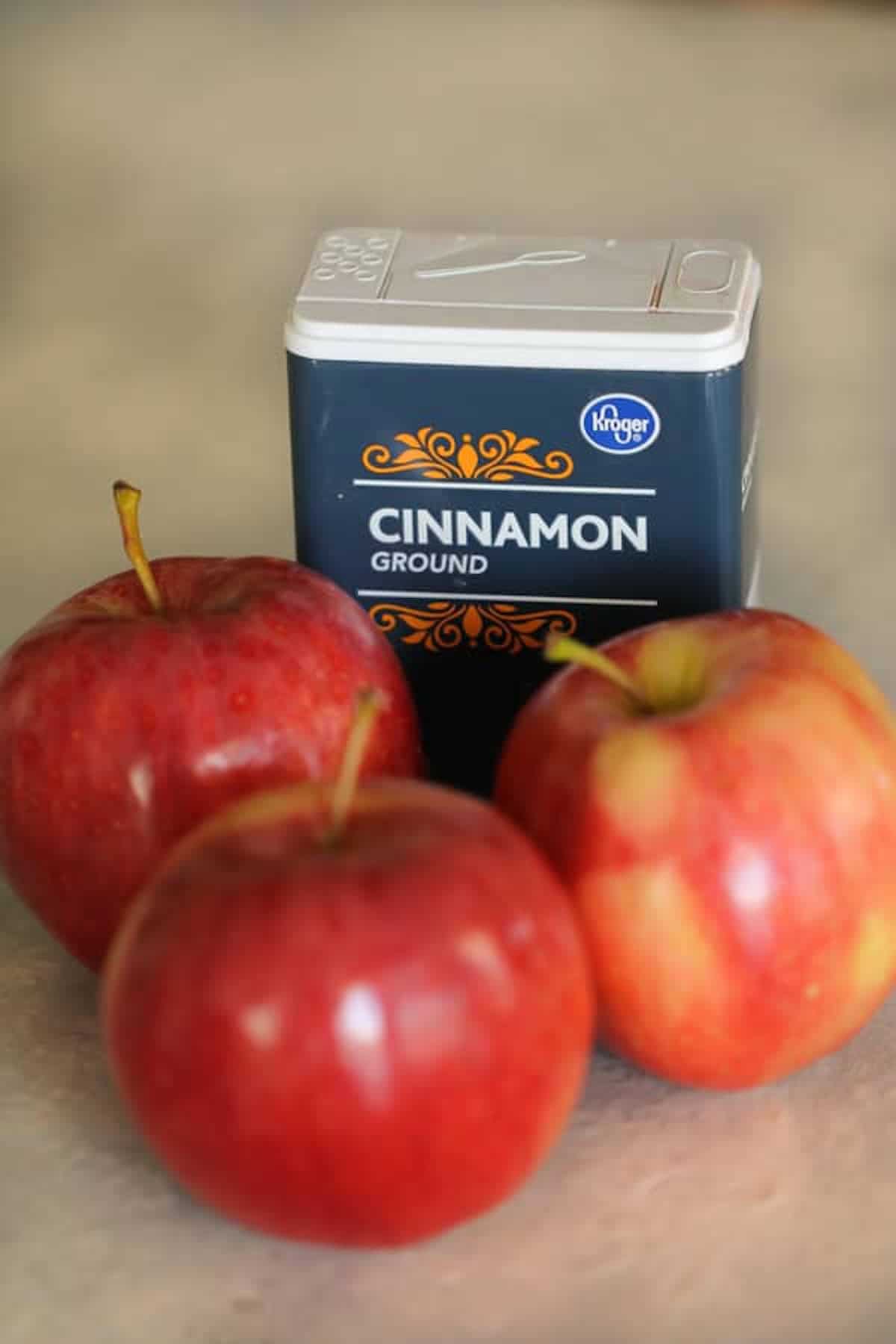 Three red apples are placed in front of a container labeled "Cinnamon Ground" on a gray surface.