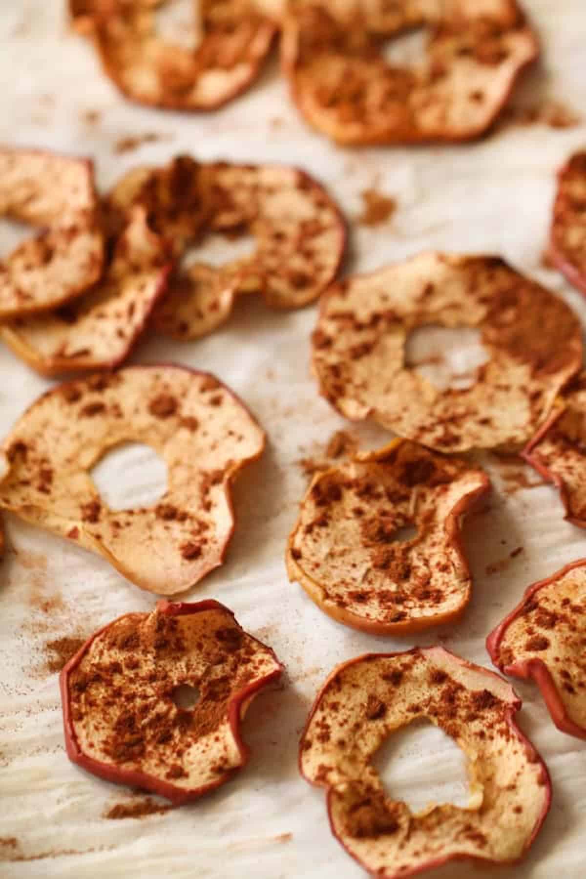 Slices of dried apple sprinkled with cinnamon are spread out on a parchment-lined surface.