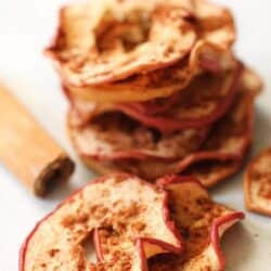 A stack of dried apple slices with cinnamon, accompanied by a cinnamon stick, placed on a white surface.