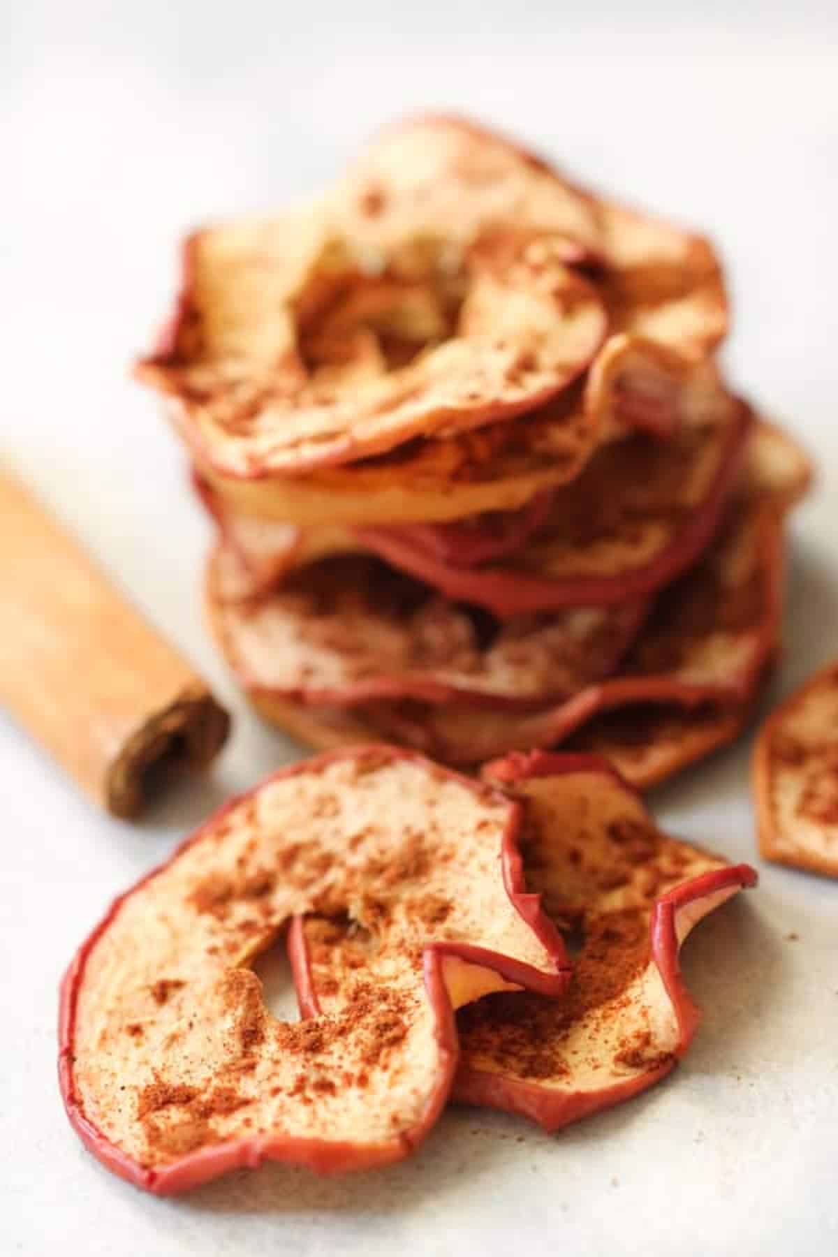 A stack of dried apple slices with cinnamon, accompanied by a cinnamon stick, placed on a white surface.