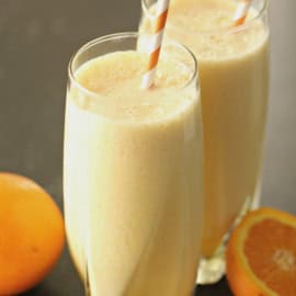 Two glasses of orange smoothies with striped straws, placed next to a whole orange and a sliced orange on a dark surface.