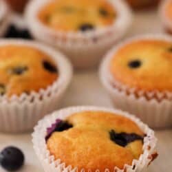 A close-up of freshly baked blueberry muffins in white paper liners, with a few scattered blueberries on a light-colored surface.