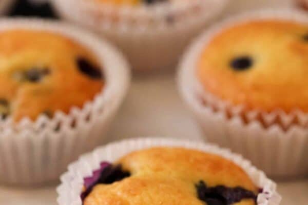 A close-up of freshly baked blueberry muffins in white paper liners, with a few scattered blueberries on a light-colored surface.