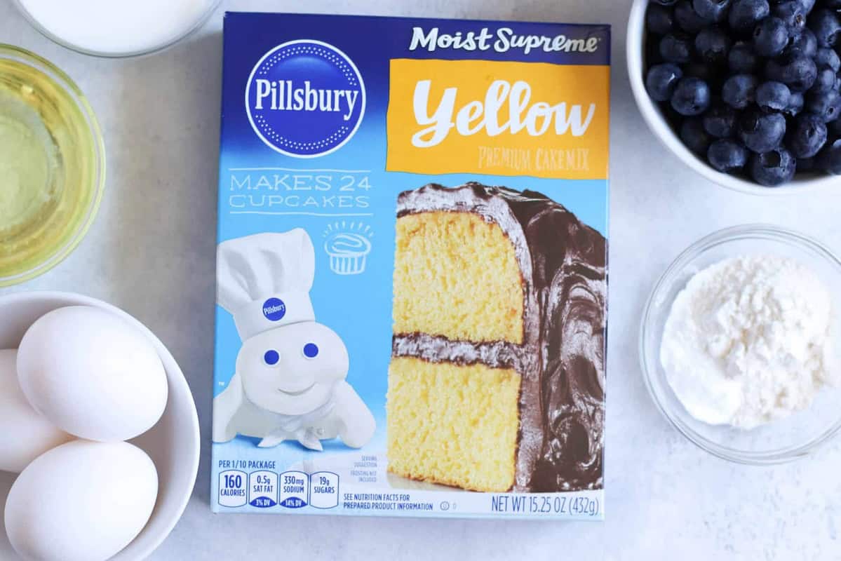 Box of Pillsbury Moist Supreme Yellow Premium Cake Mix surrounded by ingredients including eggs, oil, flour, and blueberries on a white surface.