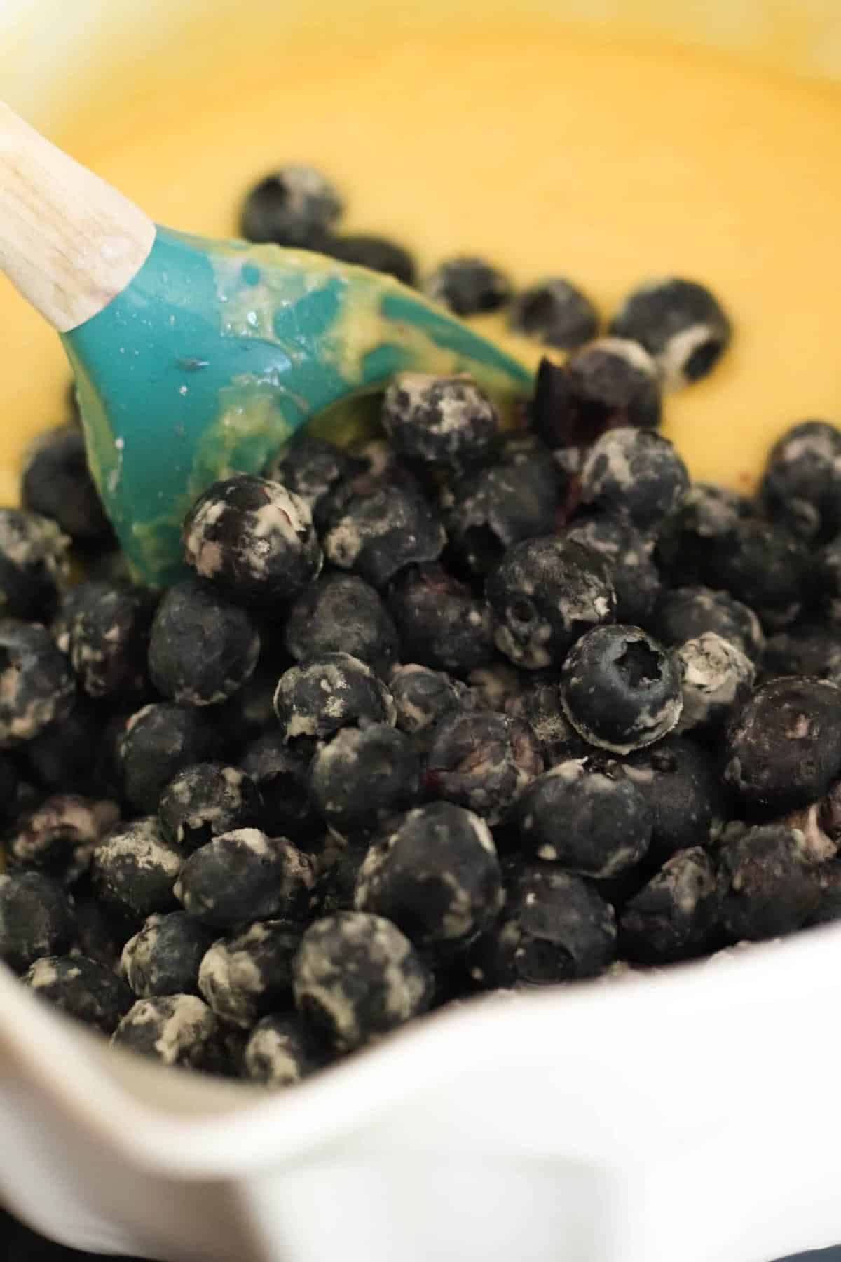 A blue spatula stirring fresh blueberries into yellow batter in a white mixing bowl.