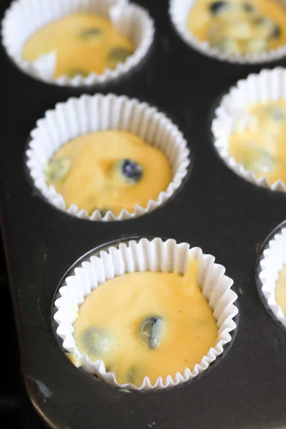 Close-up of muffin batter with visible blueberries in white paper liners placed in a metal muffin tin, ready for baking.