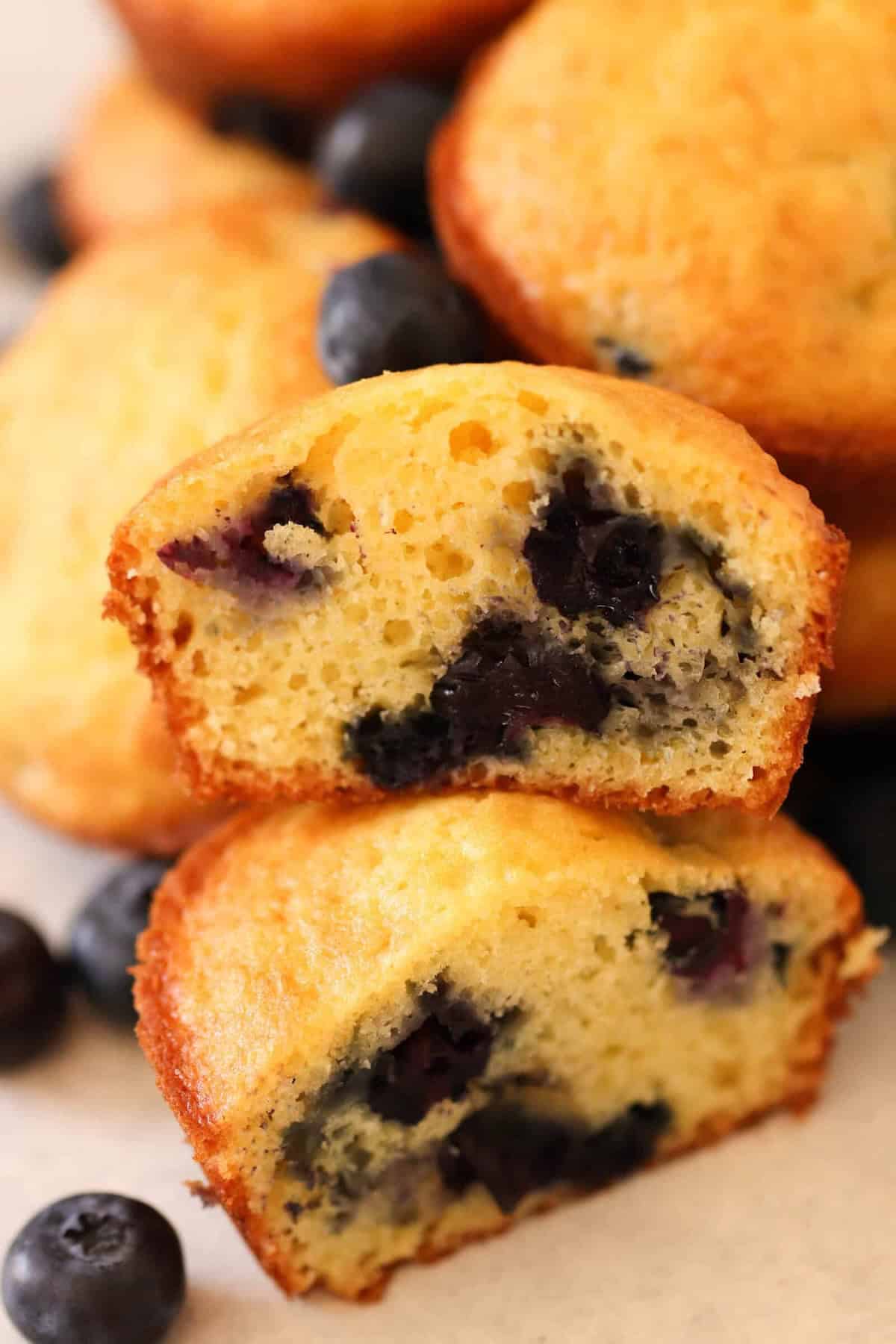 Close-up of a halved blueberry muffin with visible blueberries inside, placed in front of whole muffins and loose blueberries in the background.