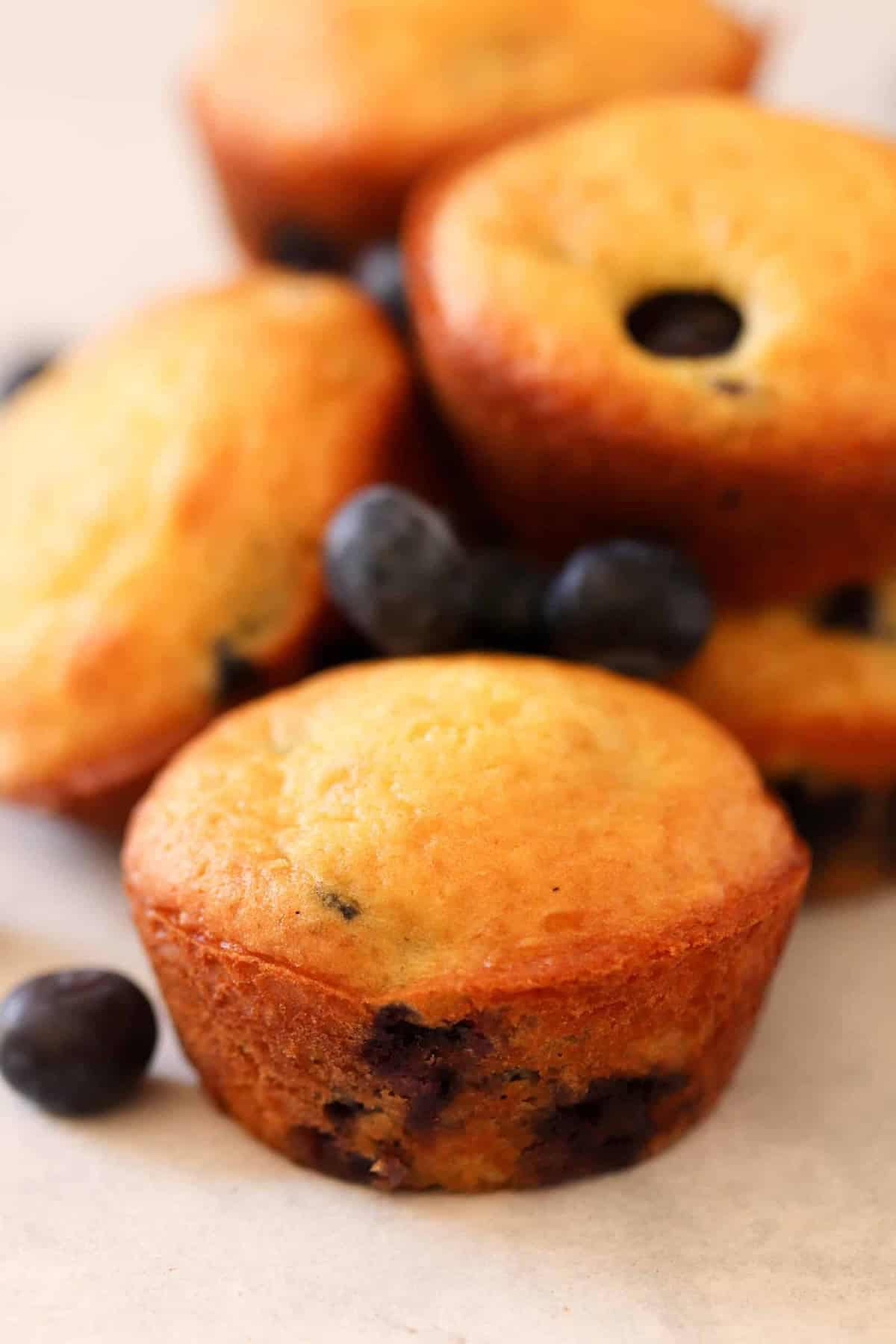 Close-up of blueberry muffins arranged on a light surface, with a few scattered blueberries around them. The muffins are golden-brown and slightly domed.