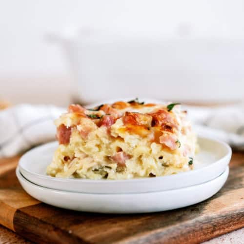 A square slice of baked hash brown casserole with visible pieces of ham, served on a white plate.