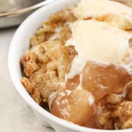 A white bowl containing a portion of apple crumble topped with a scoop of melting vanilla ice cream.