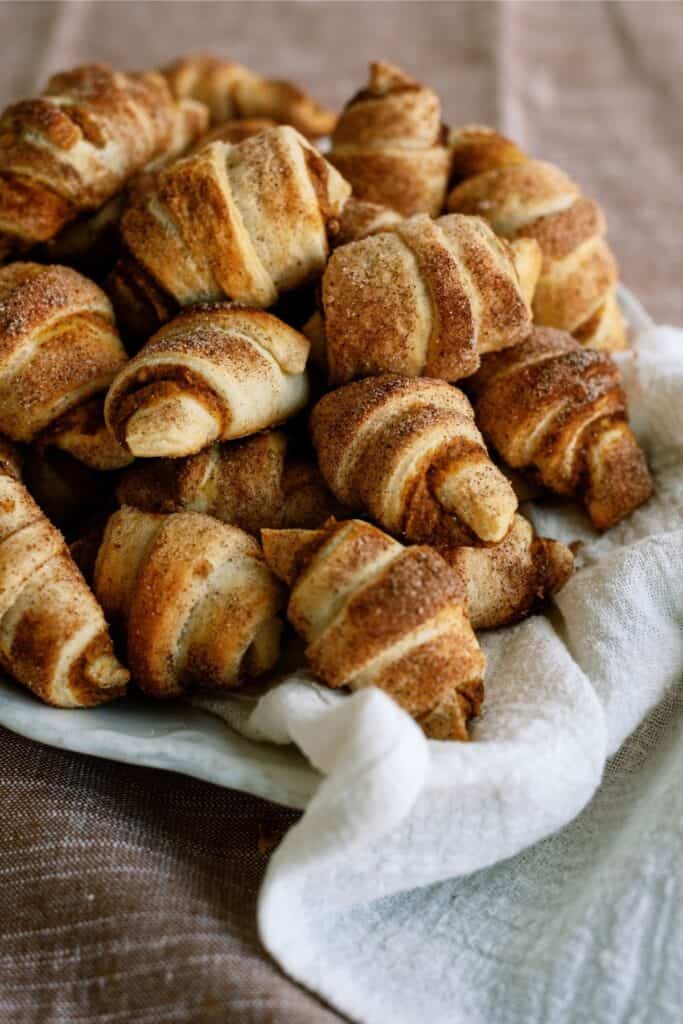 A close-up image of a pile of freshly baked croissants with a golden-brown crust, displayed on a white cloth.