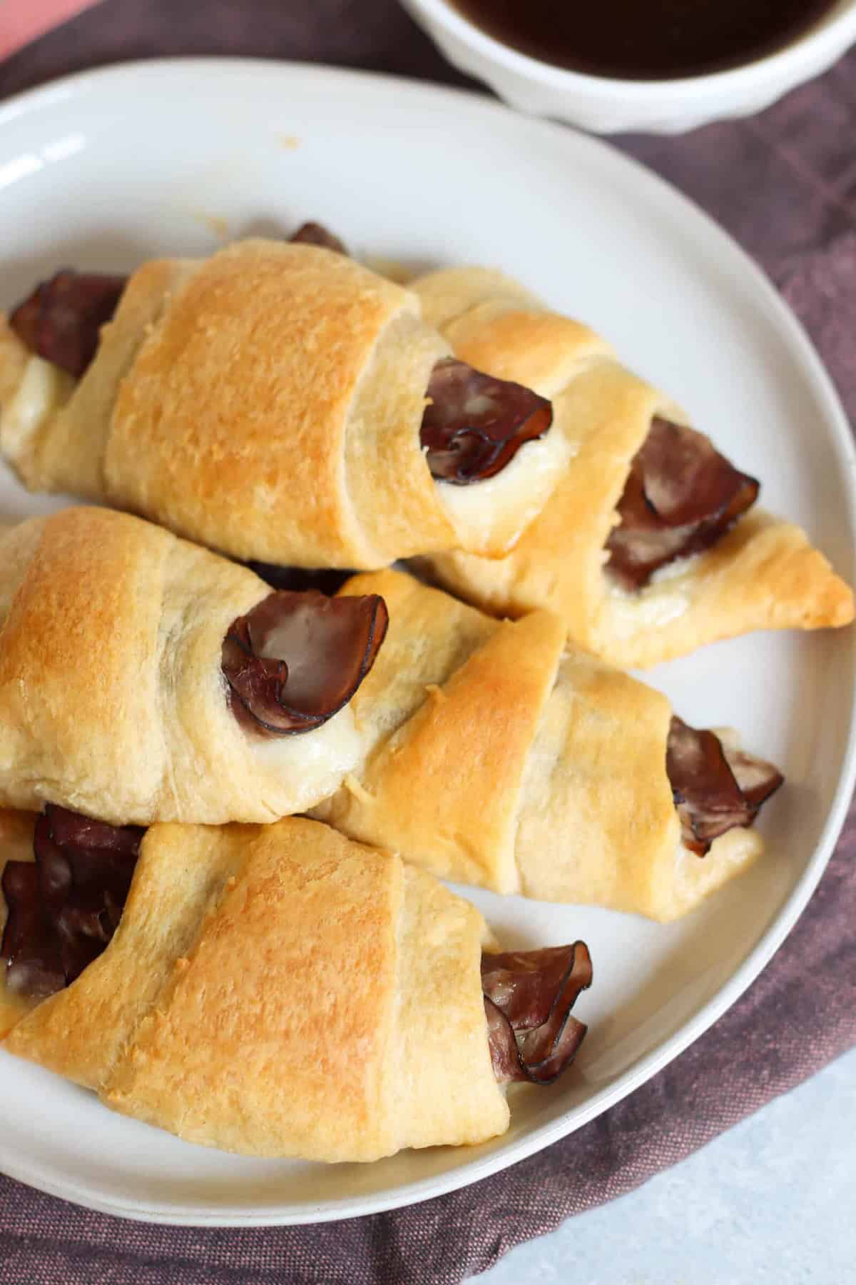 Three crescent rolls filled with slices of meat on a white plate, with a small bowl of sauce in the background.